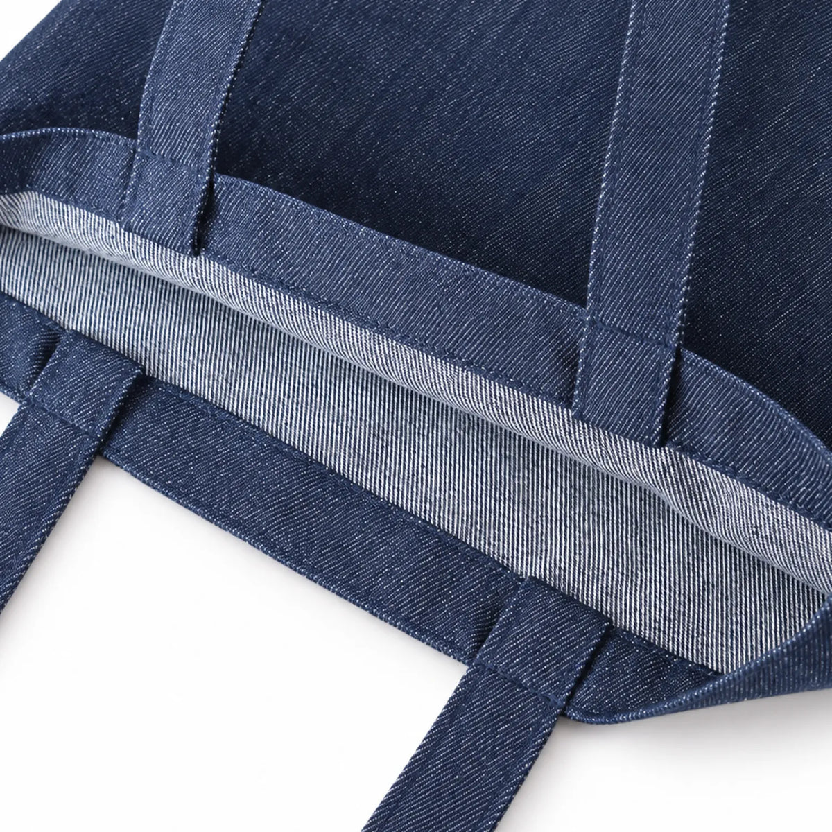 Close-up of a indigo blue denim tote bag showing the woven fabric texture, reinforced seams, and a light gray inner lining edge on a white background.