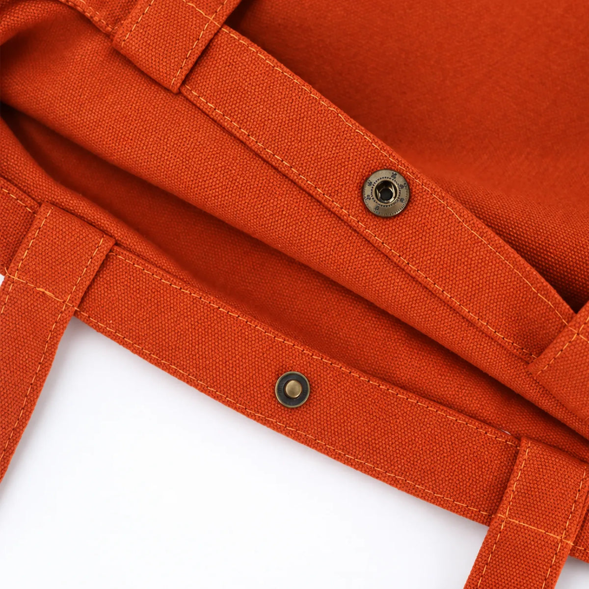 Close-up of a clay orange canvas tote bag showing the fabric texture, stitched straps, and metal snap buttons against a white background.