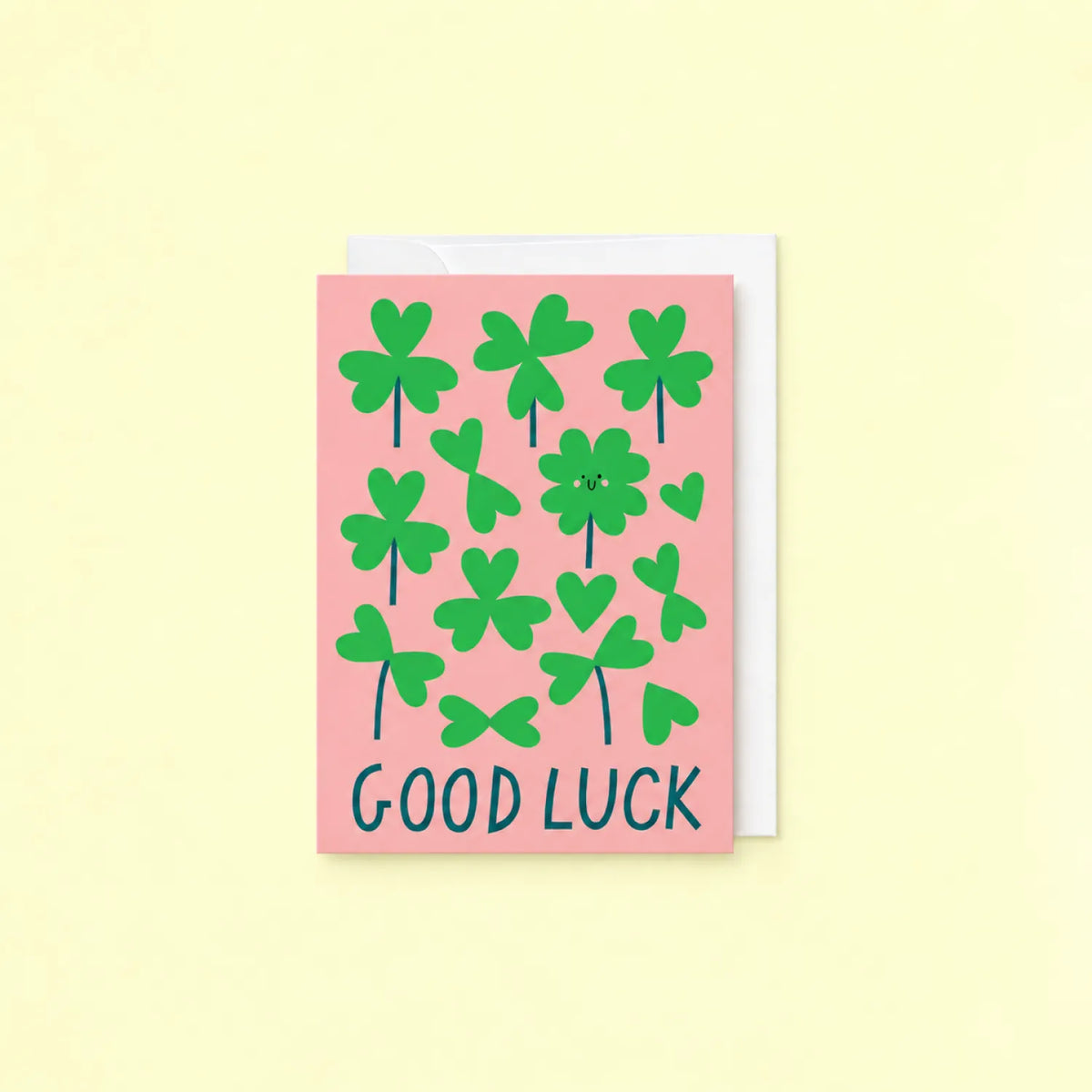 Pink Good Luck greeting card with green clovers and Good Luck lettering, shown with white envelope