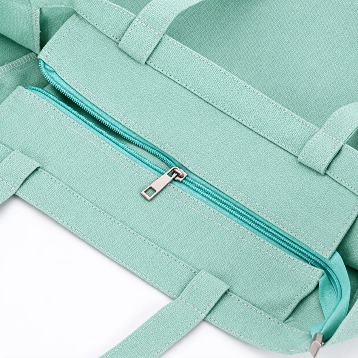 A soft teal green canvas tote bag with a matching zippered pocket and silver zipper pull, shown in a close-up detail shot against a white background.