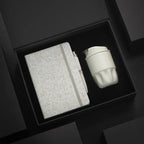 A5 cotton notebook, wheat-straw pen and white double-wall stainless steel coffee cup in open black magnetic gift box with foam insert, dramatic dark background, viewed at angle