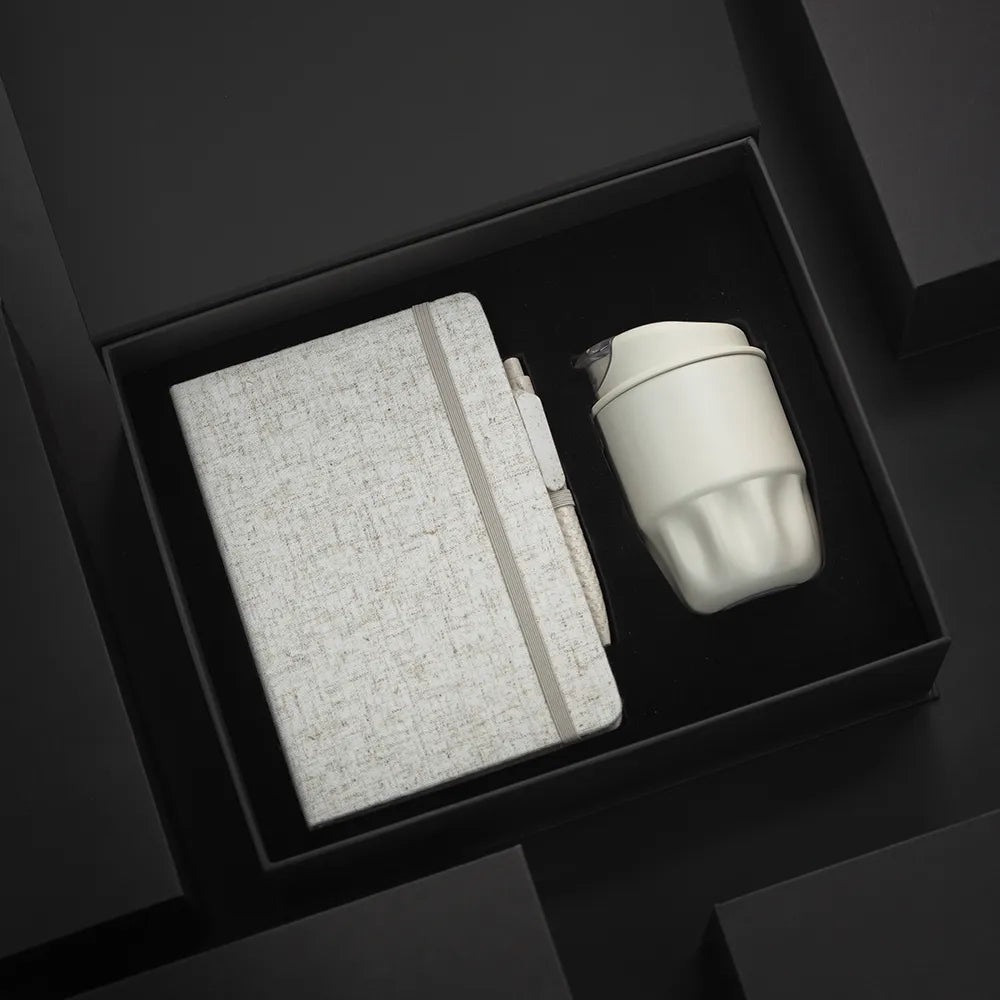 A5 cotton notebook, wheat-straw pen and white double-wall stainless steel coffee cup in open black magnetic gift box with foam insert, dramatic dark background, viewed at angle