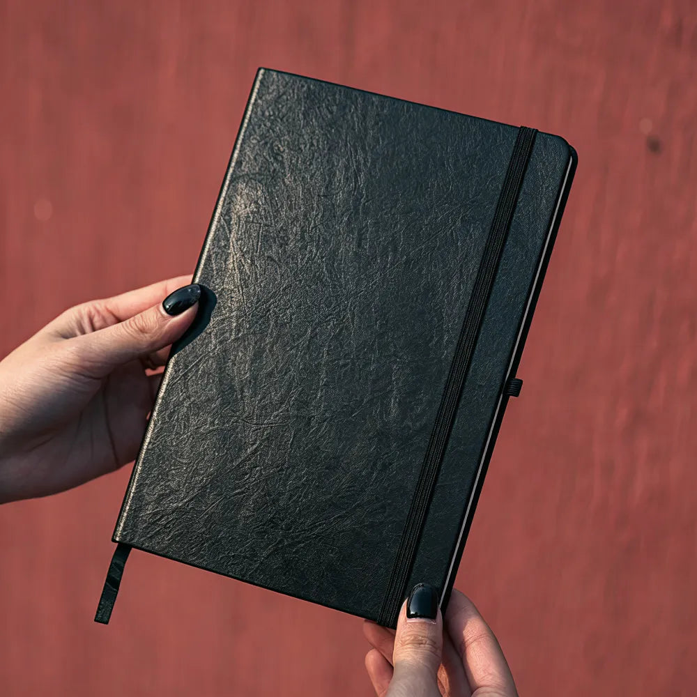 Black A5 hardback notebook with a textured leather-look cover and elastic closure band held in two hands against a warm background, ribbon marker and pen loop visible, no Wumble branding applied