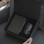 Hands opening black magnetic gift box revealing A5 Tyvek notebook and black insulated bottle in foam insert, executive employee gift set unboxing