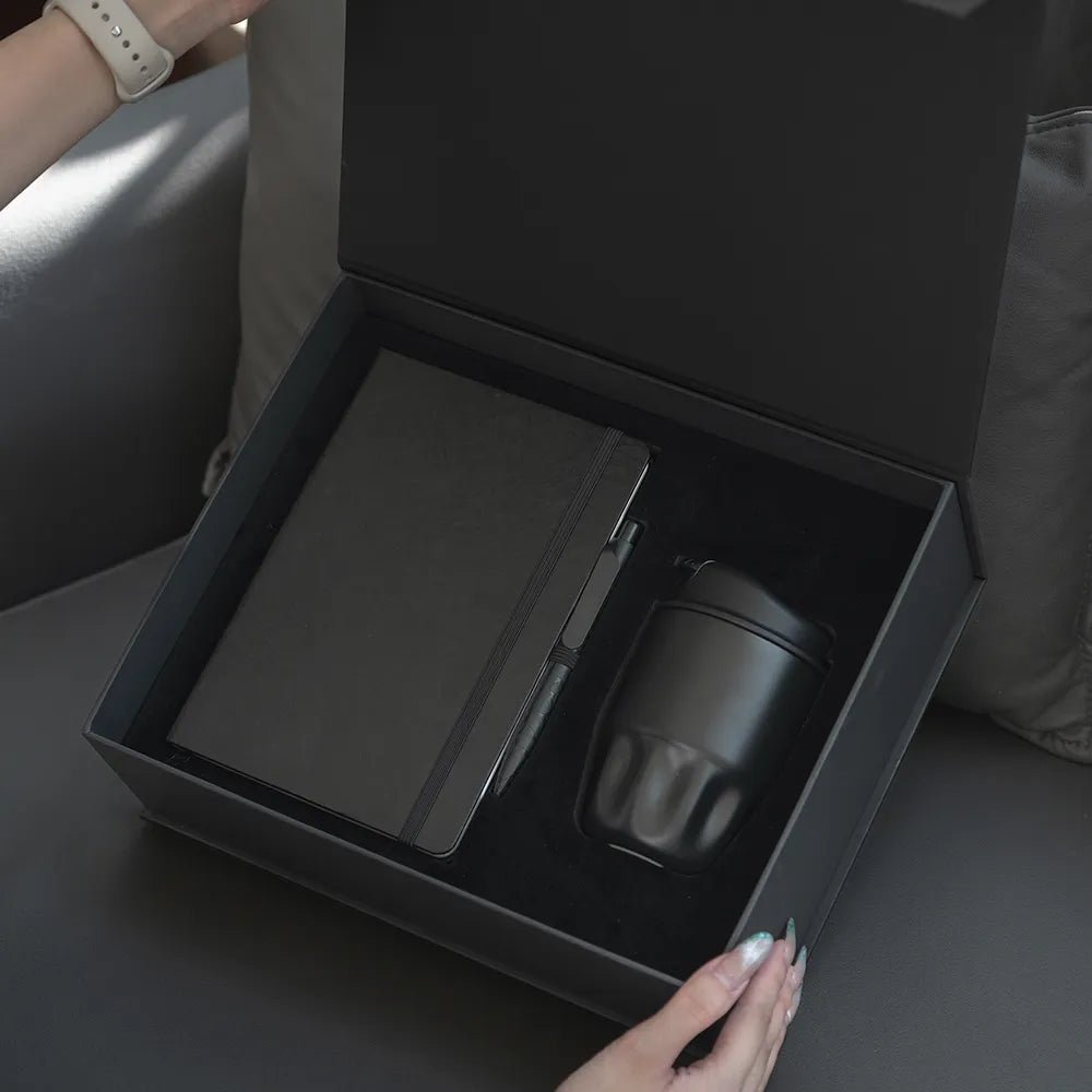 Hands opening black magnetic gift box revealing A5 Tyvek notebook and black insulated bottle in foam insert, executive employee gift set unboxing