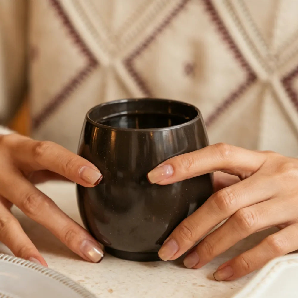 Two hands cradling a dark brown aroma recycled coffee grounds coffee cup at a table, showing the handle-free warming hold, no branding applied