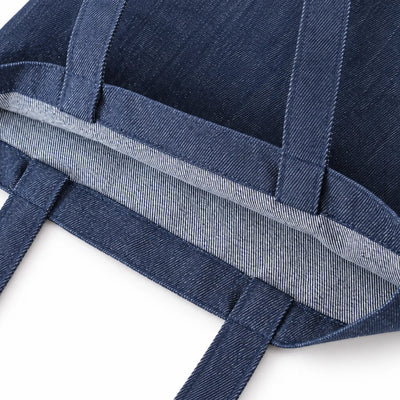 Close-up of a indigo blue denim tote bag showing the woven fabric texture, reinforced seams, and a light gray inner lining edge on a white background.