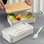 Hands lifting the bamboo lid off a wheat straw lunch box filled with salad, the lower tier holding cutlery, on a grey table, no Wumble branding applied