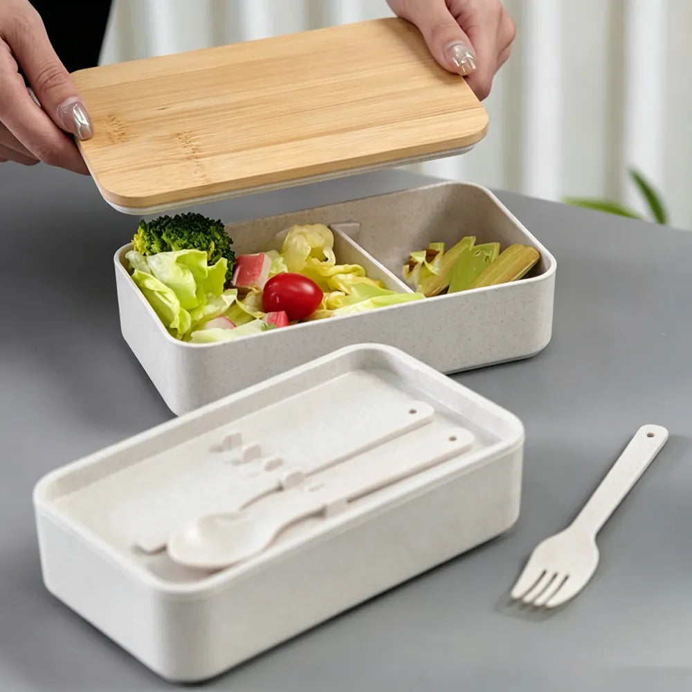 Hands lifting the bamboo lid off a wheat straw lunch box filled with salad, the lower tier holding cutlery, on a grey table, no Wumble branding applied
