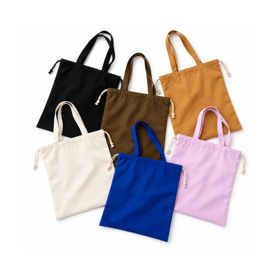 All arrange of coloured drawstring top tote bags in black, dark brown, bronze, off-white, indigo blue and pastel purple pink, overlapping flat lay against a white background. 