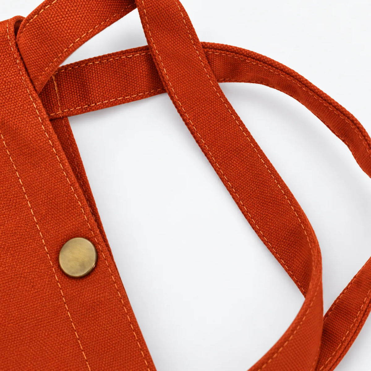 Close-up of a clay orange canvas tote bag showing the woven canvas, double strap construction, and a brass snap button closure against a white background.