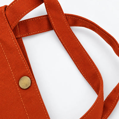 Close-up of a clay orange canvas tote bag showing the woven canvas, double strap construction, and a brass snap button closure against a white background.