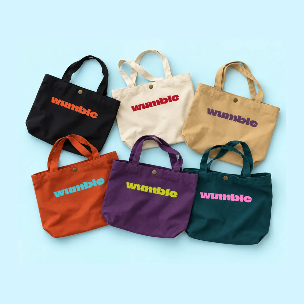 Six coloured canvas textured gusseted tote bag in black, off-white, khai, clay orange, plum purple, forest green, printed customised logo "wumble" as examples, against a white background. 