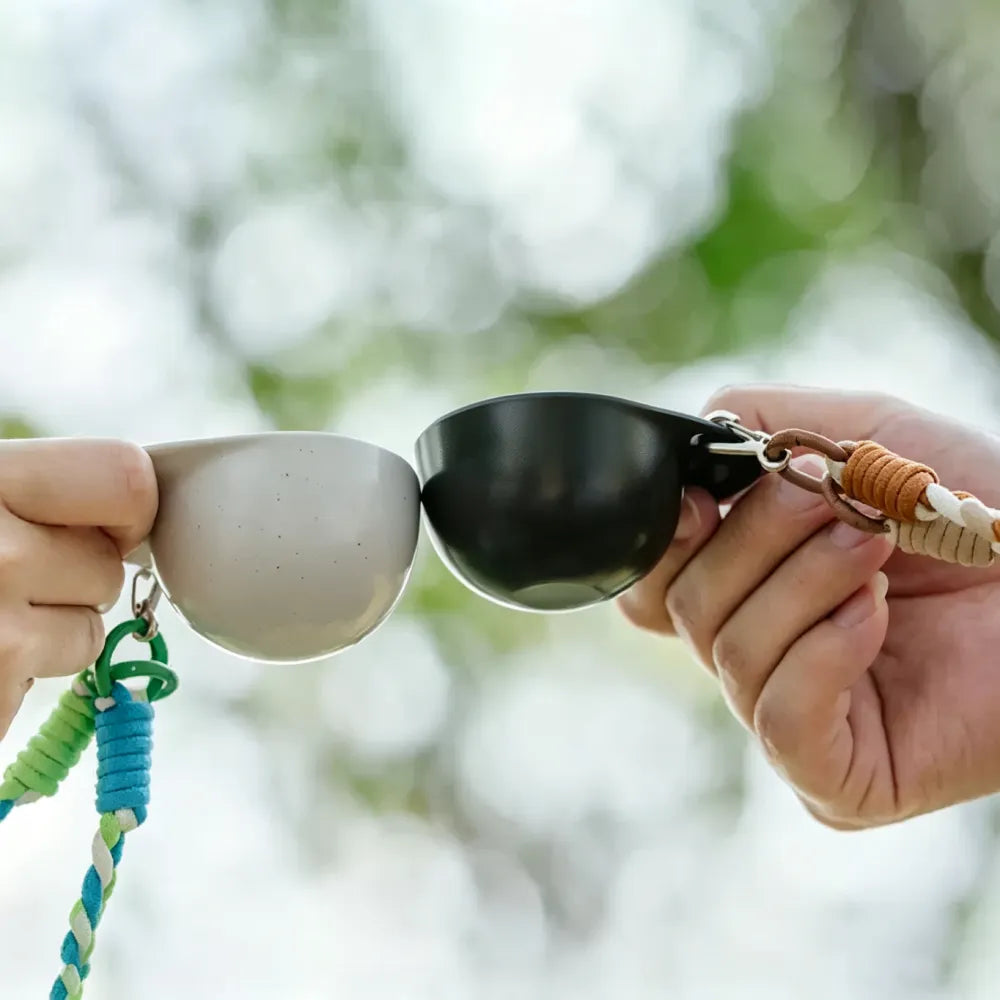 Two hands holding off-white and black hat-shaped recycled coffee grounds tasting cups on braided lanyards outdoors with green foliage behind, the cups touching