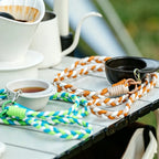 Black hat-shaped recycled coffee grounds tasting cup on an outdoor camping table beside a pour-over dripper, gooseneck kettle and braided lanyards, with an off-white cup of coffee nearby
