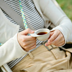 Person seated outdoors holding an off-white hat-shaped recycled coffee grounds tasting cup of black coffee by the brim, lanyard worn around the neck