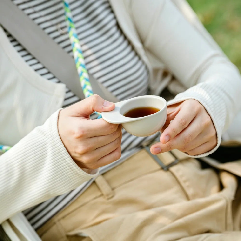 Person seated outdoors holding an off-white hat-shaped recycled coffee grounds tasting cup of black coffee by the brim, lanyard worn around the neck