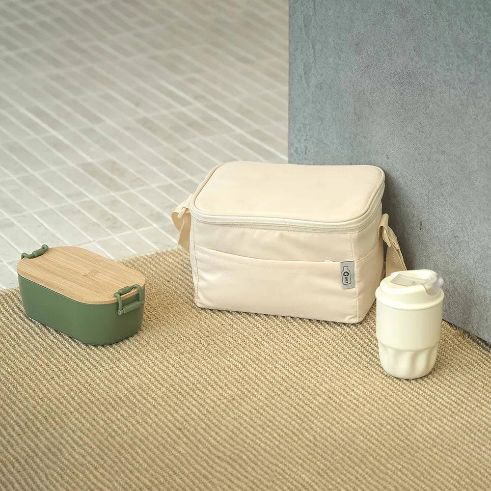 Insulated lunch bag leaning against a wall with bamboo-lid lunch box and stainless steel coffee cup arranged on a textured floor, workplace lunch break setting