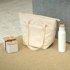Cream insulated tote bag standing upright against a wall with double-layer bamboo-lid lunch box and white insulated bottle arranged on a textured floor, everyday workplace carry kit