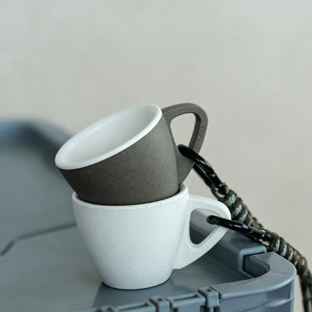 White and misty dark brown PLA espresso tasting cups nested together with a camouflage lanyard clip on a grey hard case outdoors, no branding applied