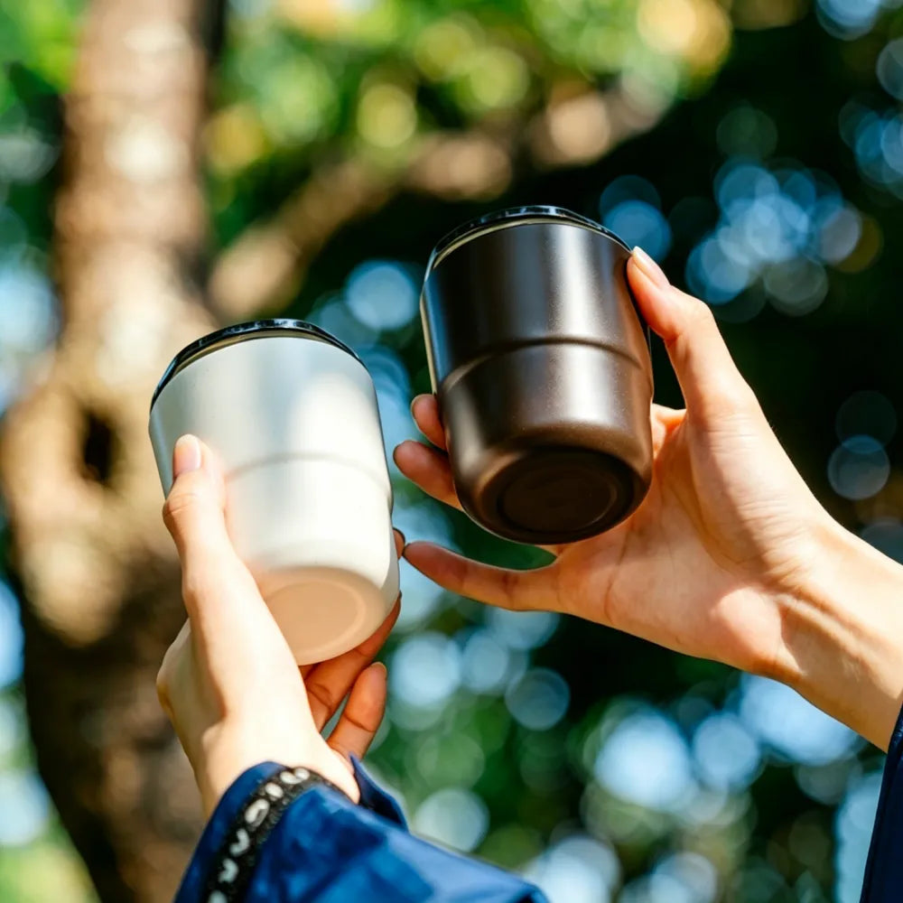 Two hands holding off-white and dark brown recycled coffee grounds everyday cups up against sunlit trees, showing both colourways, no branding applied