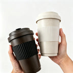 Two hands holding off-white and dark brown recycled coffee grounds travel cups side by side against a white background, showing the silicone grip bands and lids, no branding applied