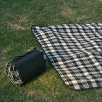Black and white check picnic mat partly unrolled on grass beside its folded carry case, showing the fleece top, no Wumble branding applied