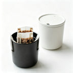 A coffee drip bag brewing inside a black reusable drip bag cup beside a lidded off-white cup on a white surface, demonstrating pour-over use