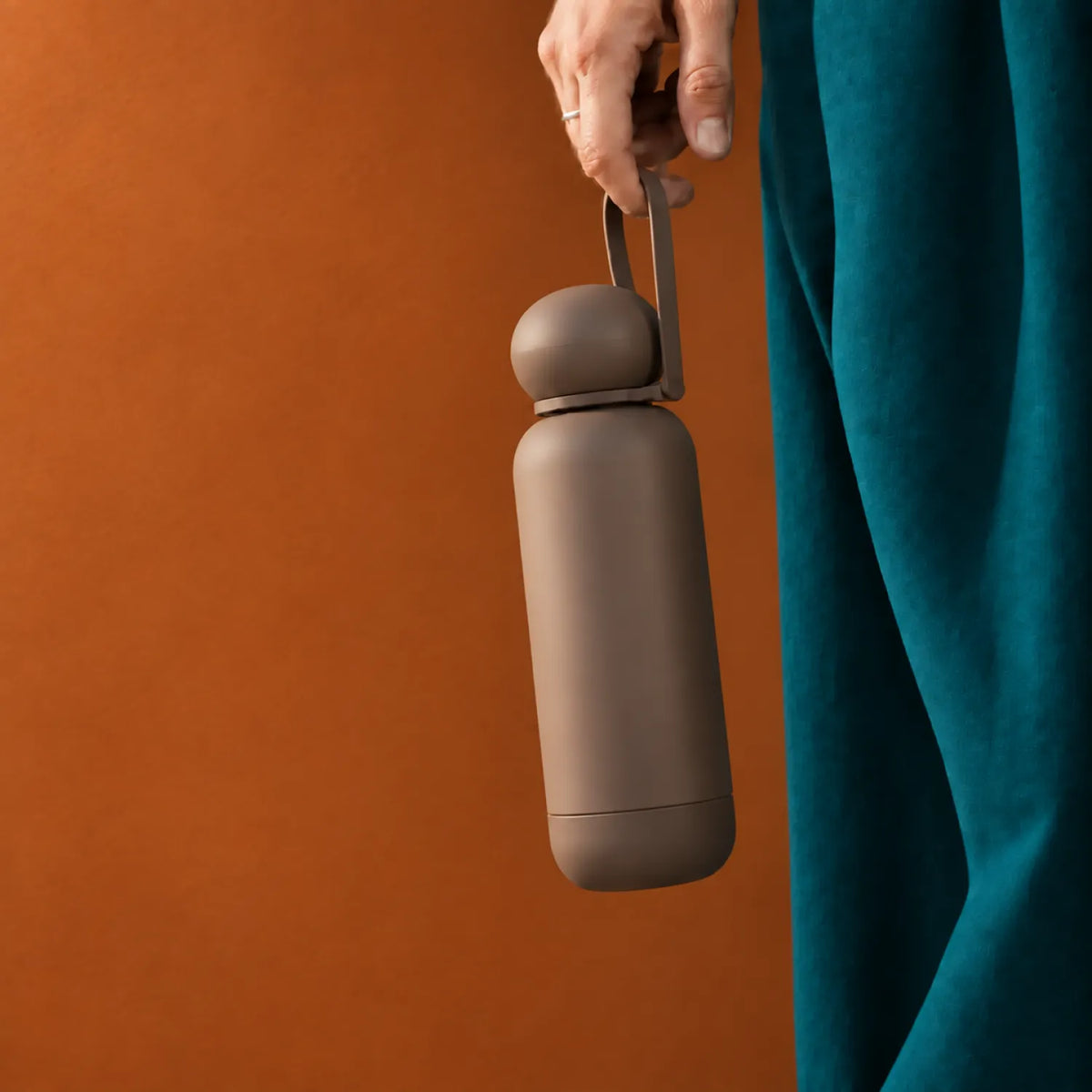 A hand holding the loop strap on a dark brown drink bottle with a rounded dome cap, richer burnt-orange ochre background.