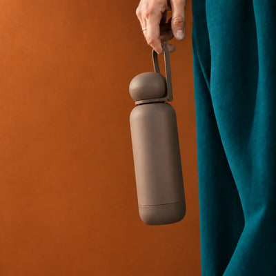 A hand holding the loop strap on a dark brown drink bottle with a rounded dome cap, richer burnt-orange ochre background.