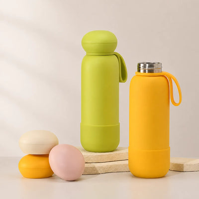 Two drink bottles, green and orange, styled with three loose caps on stone slabs against a cream backdrop.