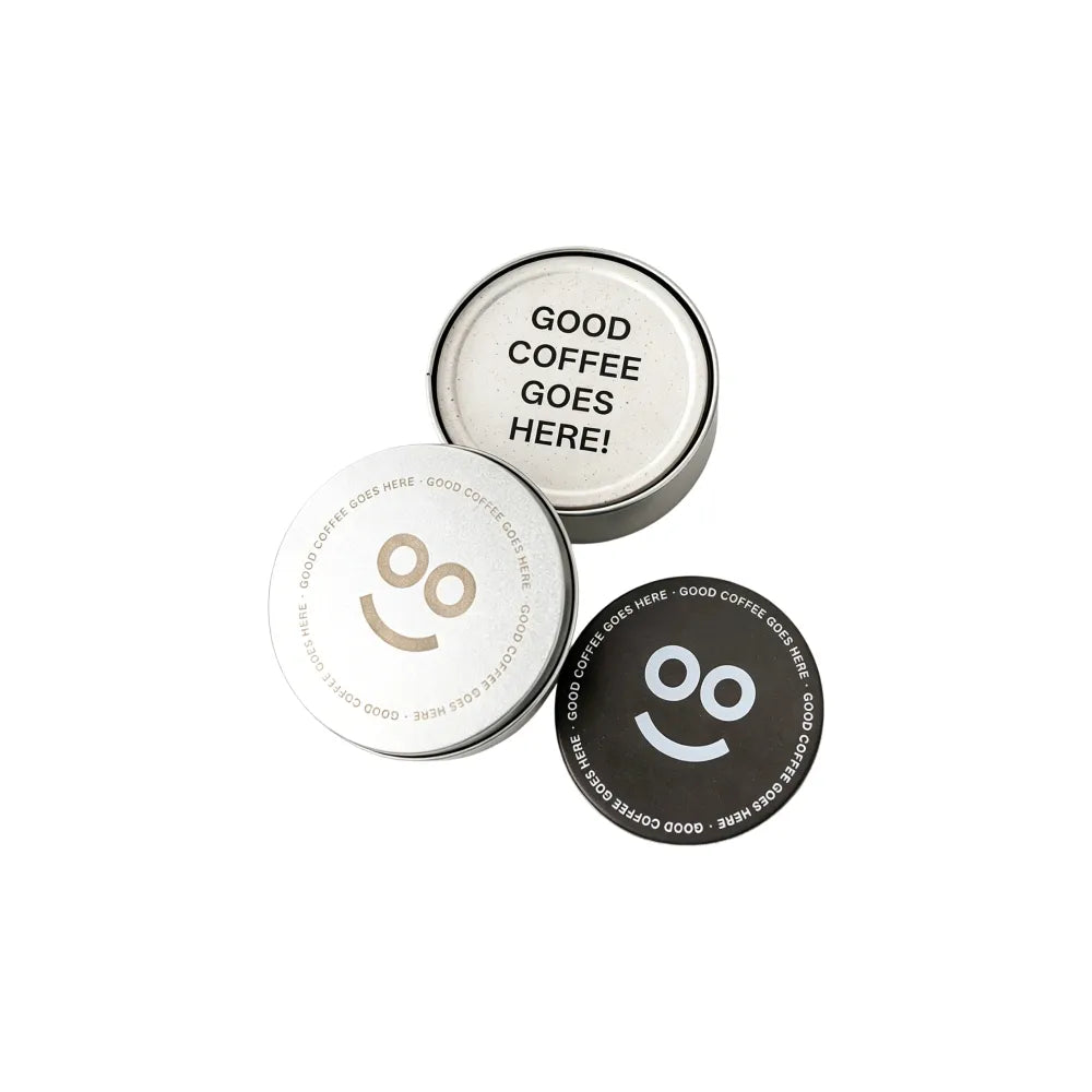 Round recycled coffee grounds coasters printed with a sample "good coffee goes here" design and smiley motif in off-white and dark brown beside a branded tin, white background, branded example