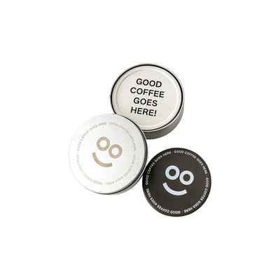 Round recycled coffee grounds coasters printed with a sample "good coffee goes here" design and smiley motif in off-white and dark brown beside a branded tin, white background, branded example