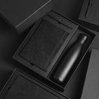 A5 Tyvek notebook, stone pen and matte black cola-style insulated bottle in open black gift box with foam insert, dramatic dark background, viewed at angle