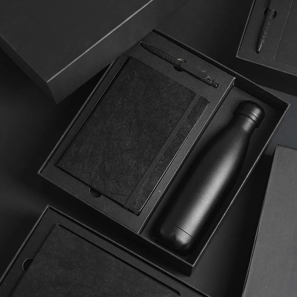 A5 Tyvek notebook, stone pen and matte black cola-style insulated bottle in open black gift box with foam insert, dramatic dark background, viewed at angle