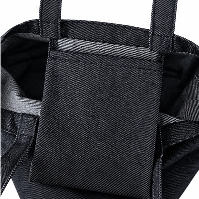 Close-up of a black denim tote bag showing a front slip pocket and the textured woven fabric of the main body.