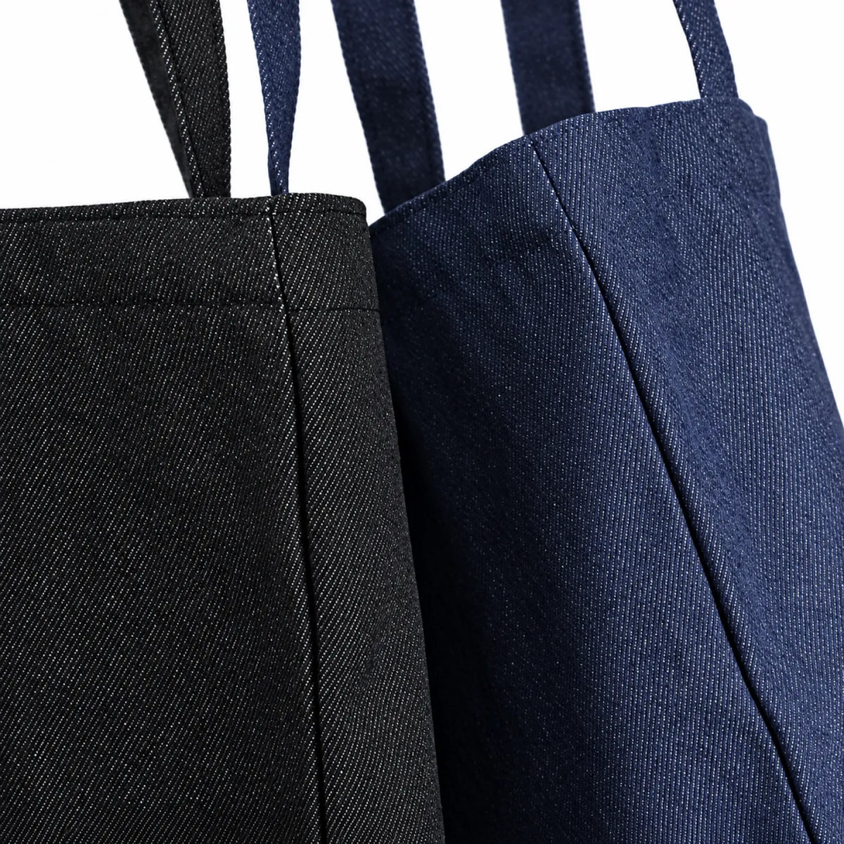 Two denim tote bags shown in a close-up studio shot, one in black and one in dark indigo blue, both with matching shoulder straps against a white background.