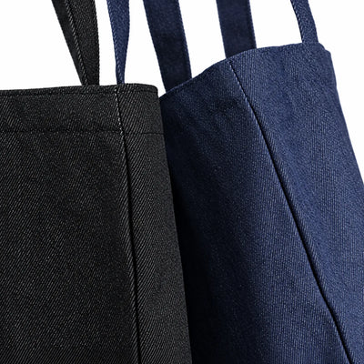 Two denim tote bags shown in a close-up studio shot, one in black and one in dark indigo blue, both with matching shoulder straps against a white background.