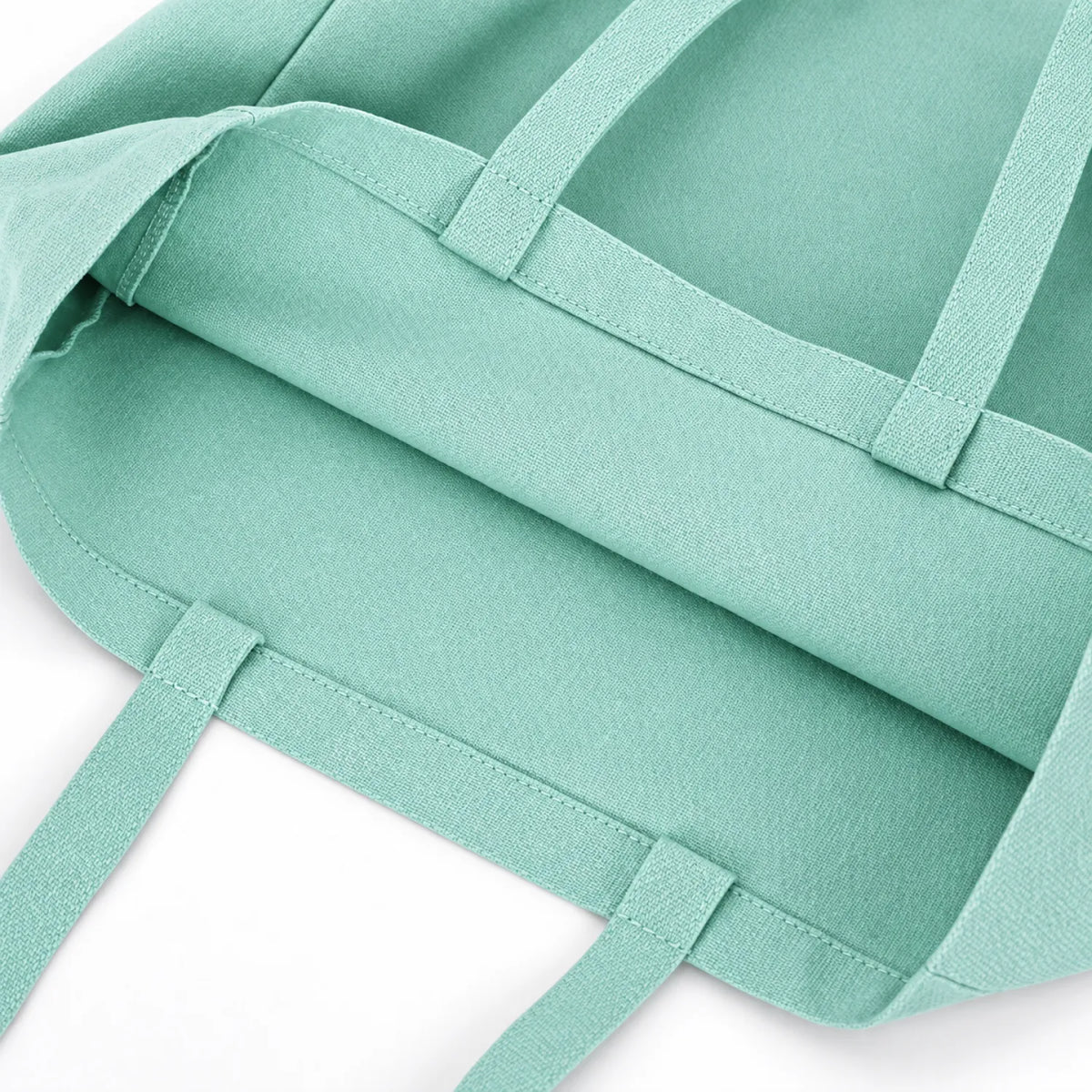 A close-up of a soft teal green canvas tote bag showing its fabric texture, reinforced stitching, and webbing handles against a clean white background.