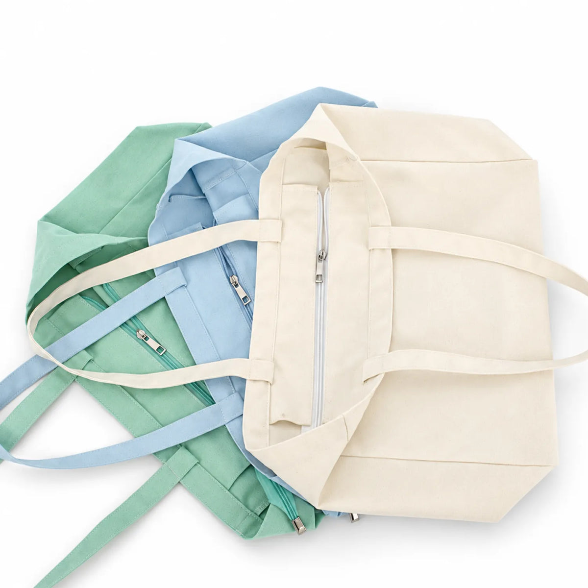 Three canvas zippered tote bags in off-white, soft blue, and soft teal green, arranged overlapping on a white background.